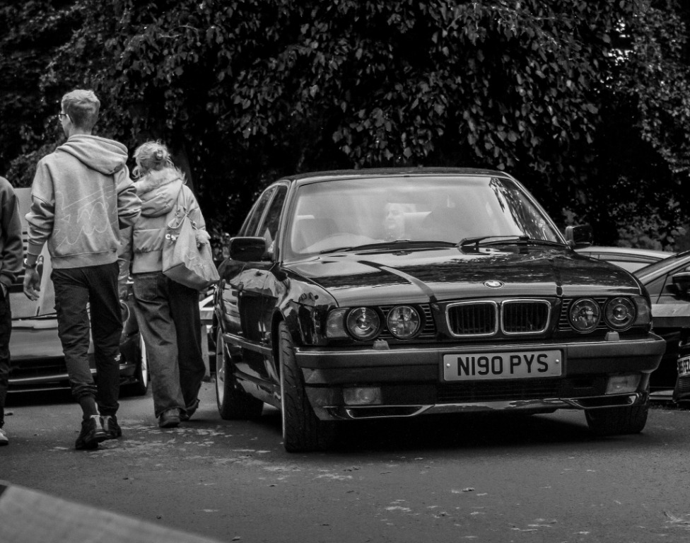 Monochrome BMW coupe parked beside pedestrians on a city street.