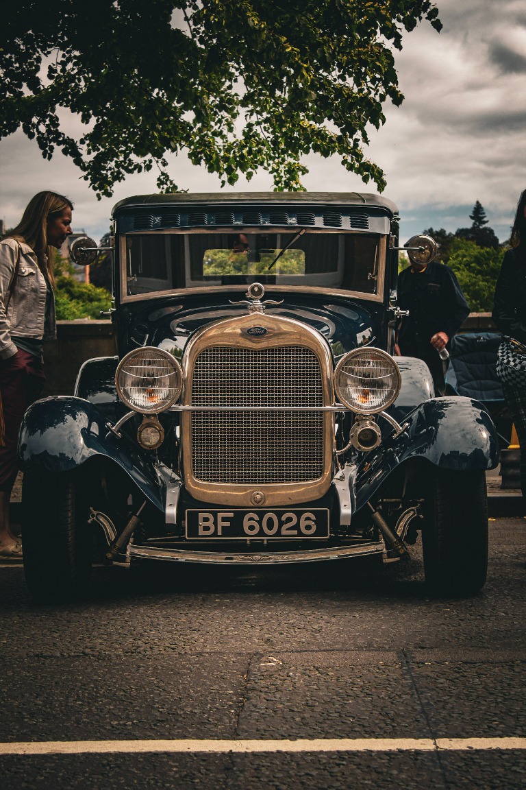Front view of a classic Ford car parked beside a tree-lined road.