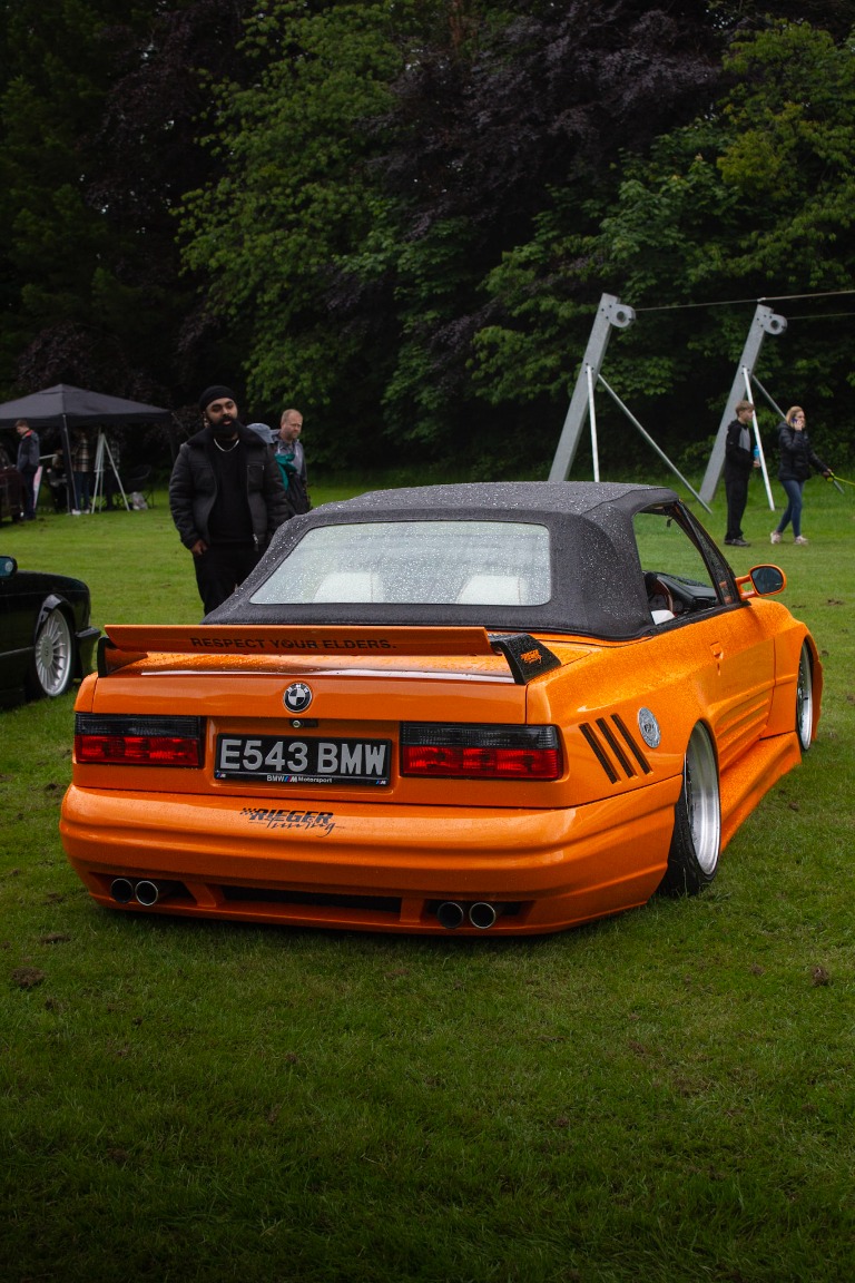 A bright orange BMW roadster at a classic car show in the rain.