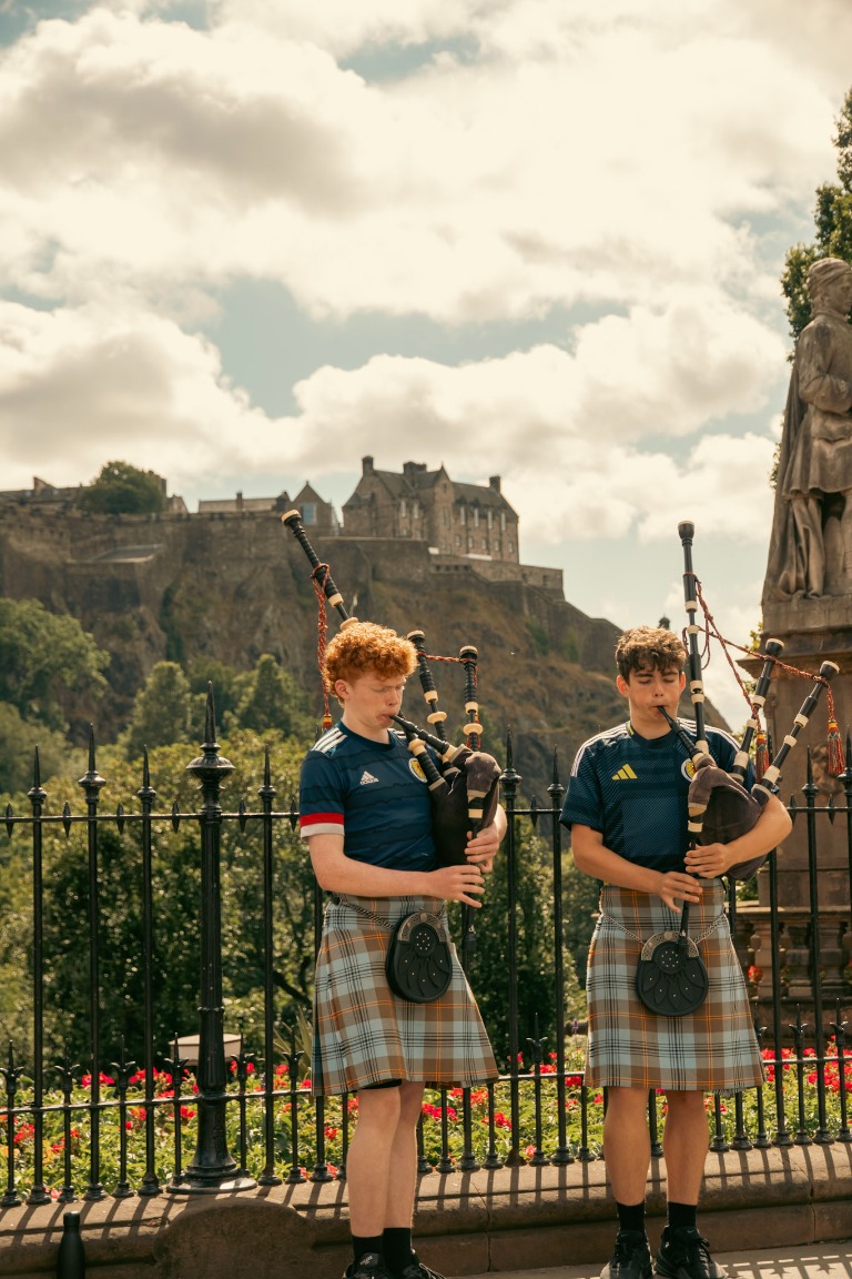Two young bagpipers play outdoors beside a historic castle.