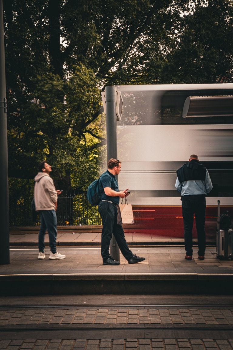 Three commuters waiting beside a bus beneath leafy trees.